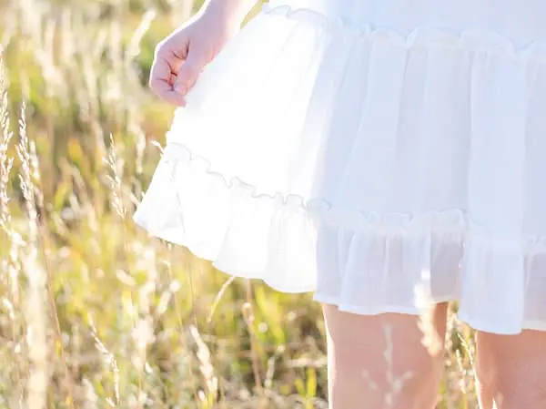 Close-up of a person gently touching the hem of a white tiered dress with ruffles, standing in a golden, sun-drenched grassy meadow.