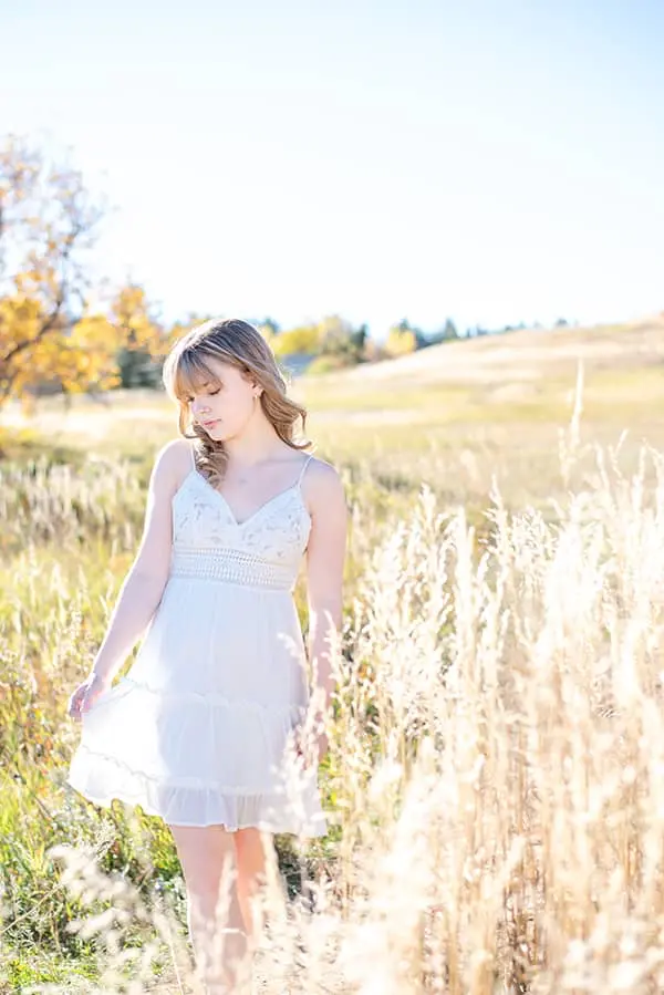 Young woman in a white lace dress looking down thoughtfully while standing in a sunlit grassy field during golden hour