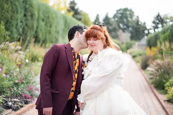 Groom in a burgundy suit kissing the bride's cheek on a paved pathway lined with garden flowers and trees