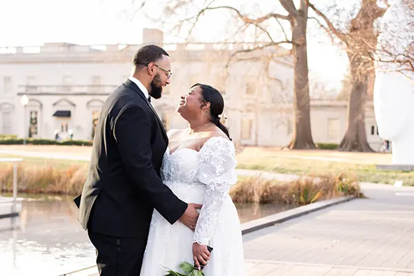 Bride in a white lace gown holding a bouquet while looking at her groom in a tuxedo near an estate pond
